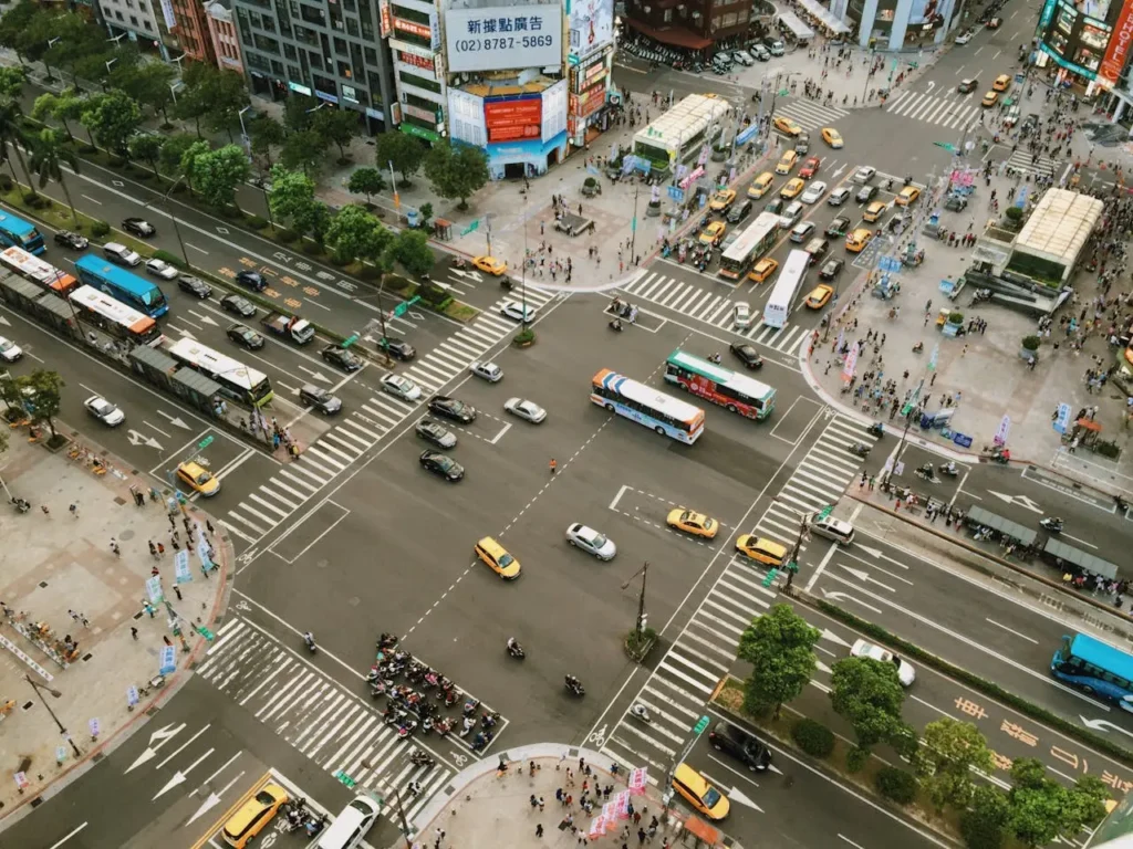 Aerial view of a busy city intersection packed with cars, buses, taxis, motorbikes, and pedestrians crossing in multiple directions.