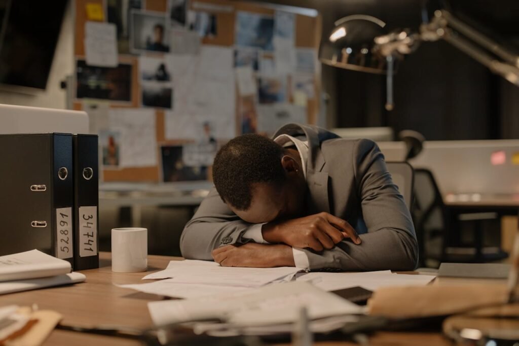 Exhausted man in a business suit resting his head on a cluttered office desk, surrounded by files and paperwork.

