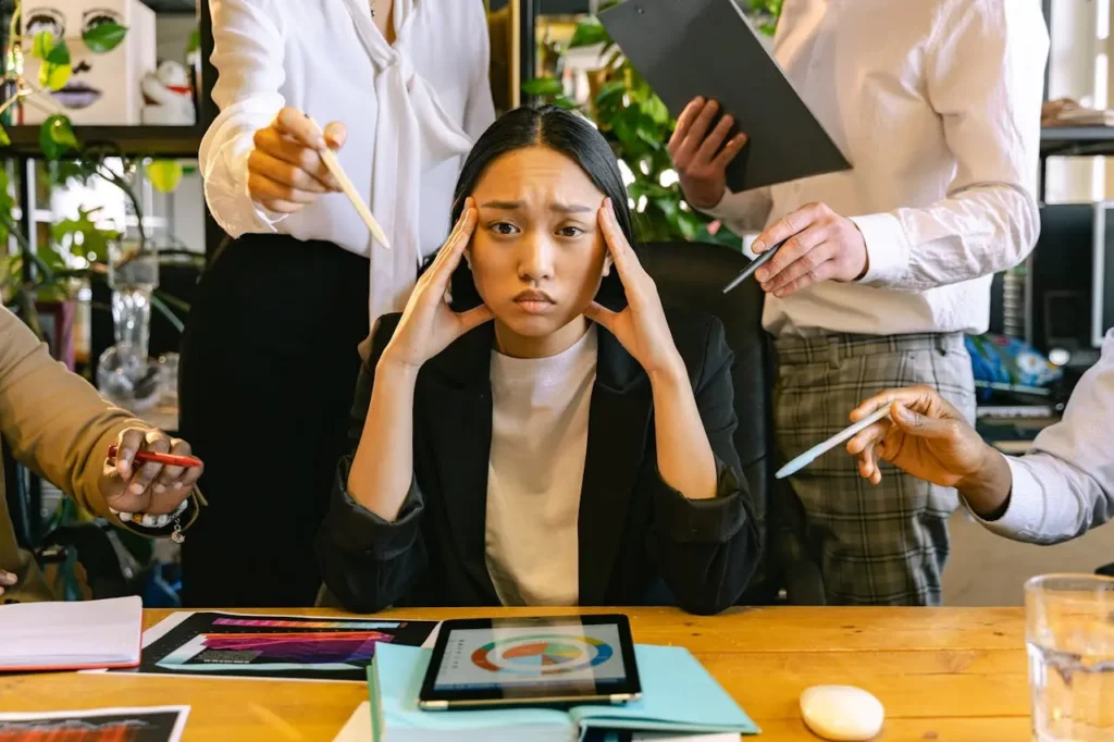 Stressed woman at a desk holding her head while surrounded by coworkers demanding attention, pointing pens and papers at her.