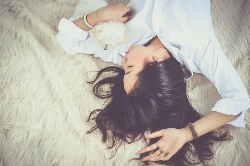 A young woman peacefully napping on a soft, cream-colored rug, gently holding a fluffy white puppy close to her heart.